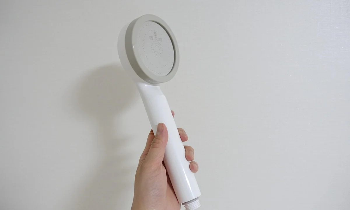 A hand holding a white handheld shower head against a plain light-colored wall.