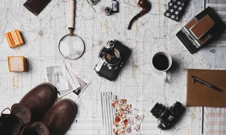 Top-down view of a map spread on a table with travel gear scattered across it—boots, camera, compass, magnifying glass, notebook, and coffee cup—evoking fieldwork in tarantula biogeography.