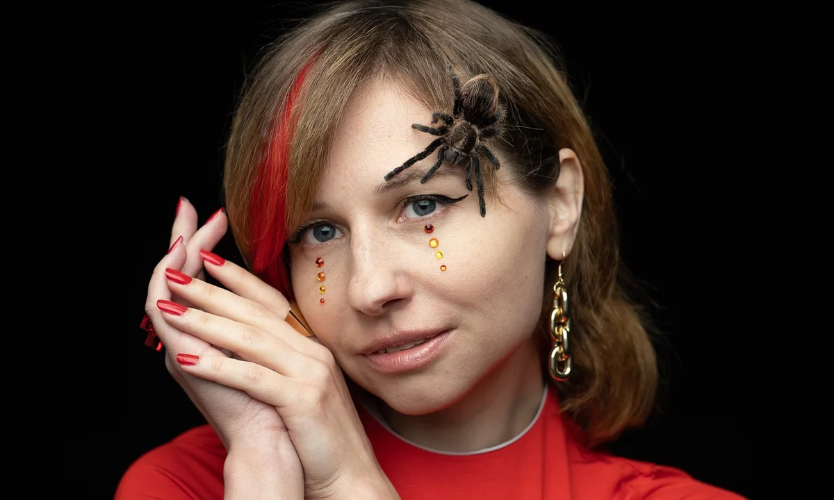 A woman wearing a red outfit with a tarantula perched on her head, against a dark background.