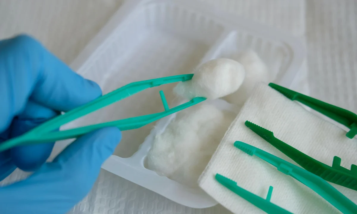 Close-up of a gloved hand using teal forceps to handle a white bolus in a small tray, with additional teal tools laid out nearby.