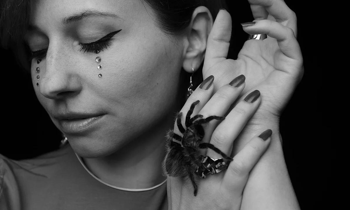 Black-and-white portrait of a woman with winged eyeliner and teardrop makeup, a tarantula resting on her hand.