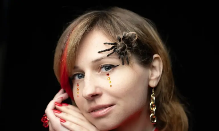 Portrait of a woman with a tarantula perched on her forehead, wearing makeup and jewelry.
